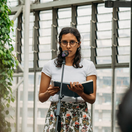 Alex Chand, an Asian/multiethnic woman with shoulder-length dark hair and glasses, wears a white tee and floral skirt. She stands in front of a bright window and holds a book. She is giving a public reading, speaking into a microphone.