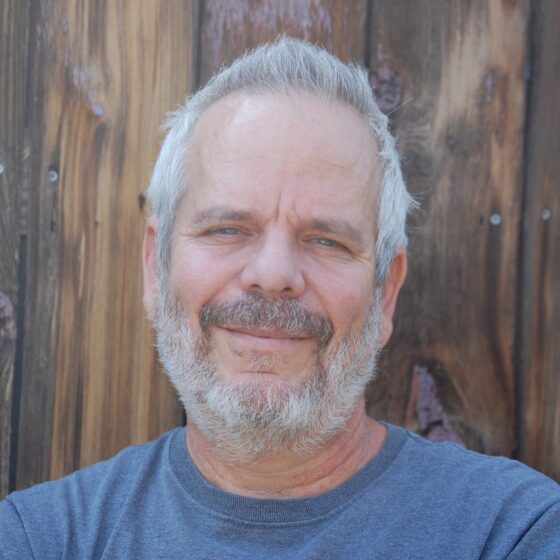 Gabriel Arquilevich, a white man with white hair and gray beard, wears a blue tee shirt and smiles in front of a wooden fence.