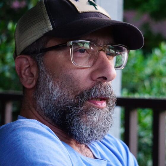 Nicholas Regiacorte, a white man with salt-and-pepper beard wearing a brown trucker cap and clear-framed glasses, appears to be sitting on a porch, looking away from the camera.