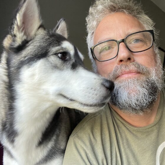 Shane Seely, a white man with gray beard and black-framed glasses, poses next to a Husky that appears to be about to lick him. It looks off to the right.