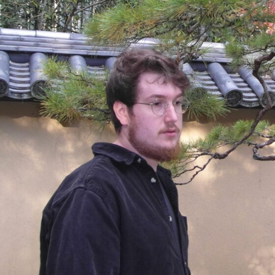 Ian Cappelli, a white man with brown hair and beard and glasses, wears a black pullover. He stands in front of a Japanese-style roofed building, next to a thin tree.