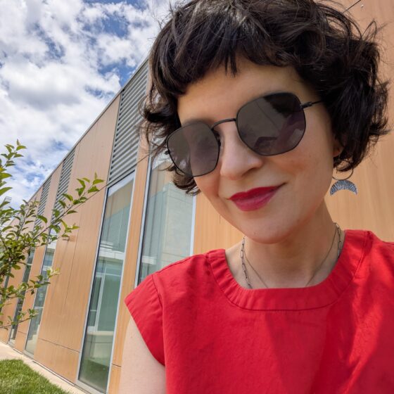 A woman with short dark hair and big sunglasses. She's wearing a red blouse and lipstick and standing in front of a brick and glass building outside