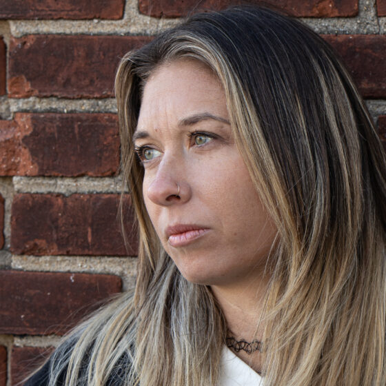 Moriah Cohen, a white woman with long highlighted hair and a silver nose ring, stands in front of a brick wall and looks into the distance.