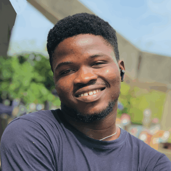 Adeniyi Odukoya, a young Nigerian man with short hair, smiles broadly. He wears a black tee shirt and is standing outdoors in a garden-type setting.