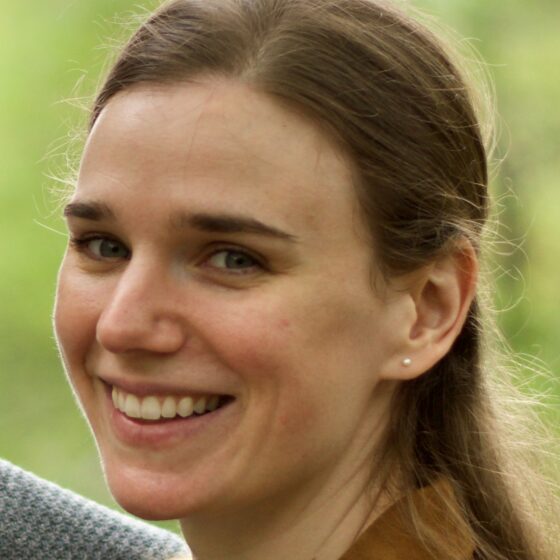 Katherine Damm, a white woman with brown hair half-pulled-back, smiles broadly in front of a background of greenery.