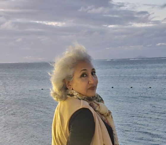 Shikhandin, an older Asian woman with white hair, is elegantly dressed in a vest and scarf in front of a large body of water.