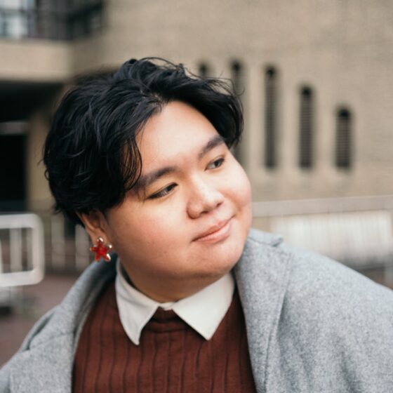 Conan Tan, an Asian person, wears a sparkly star earring, gray coat, brown sweater, and white collar poking out underneath. They smile and look into the distance. Behind them are some gray buildings.