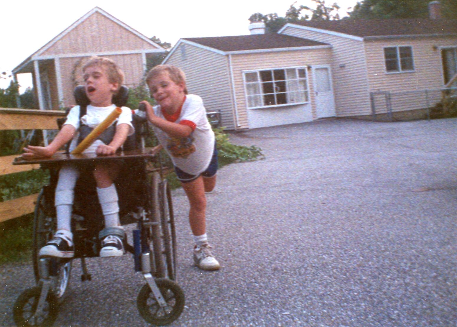 A young Brian and Danny Trapp, perhaps 5 to 7 years old, playing in their driveway in the mid-80s. Brian is pushing Danny's wheelchair and both are caught up in the fun of the game. A beige ranch house is in the background.