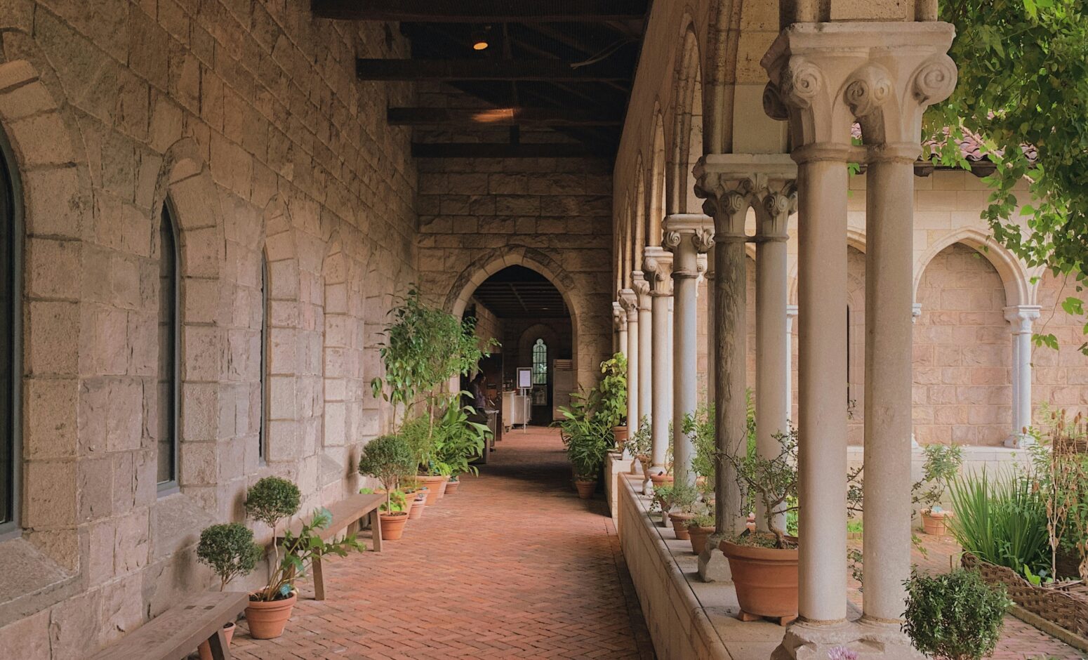 Covered walkway at the Met Cloisters, supported on one side by columns. There are potted trees alongside the wall. At the end of the walkway is an arched doorway.