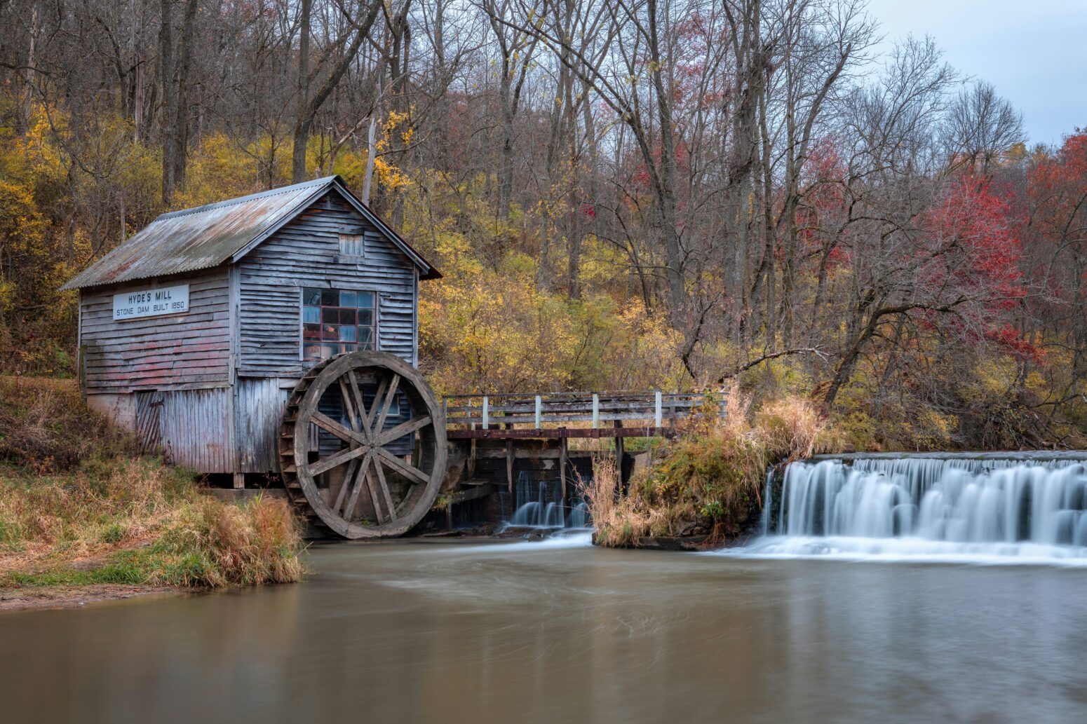 A 19th-century water mill, made of brown wood with a water wheel, sits next to a dam. It's autumn and the leaves are turning. The sign on the mill reads "Hyde's Mill--stone dam built 1850."
