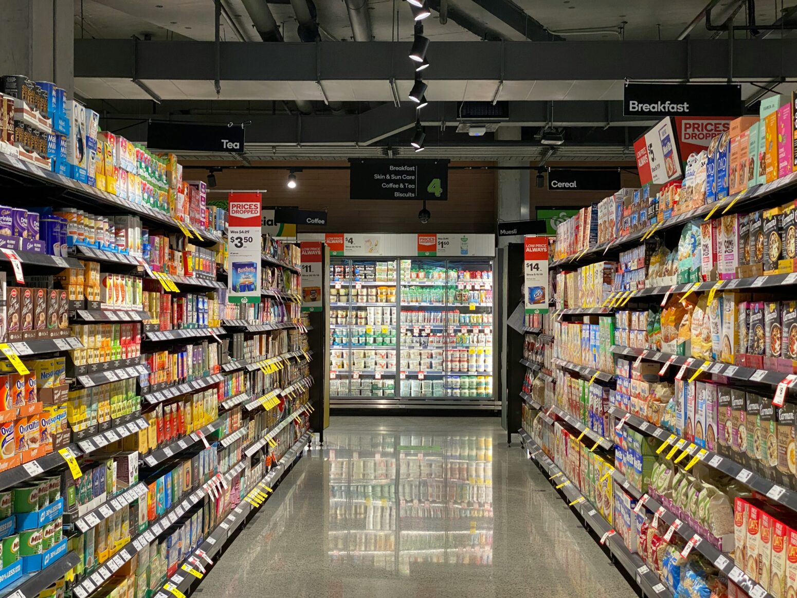 Interior of a grocery store, looking down an aisle of brightly colored packaged goods at a glowing refrigerator.