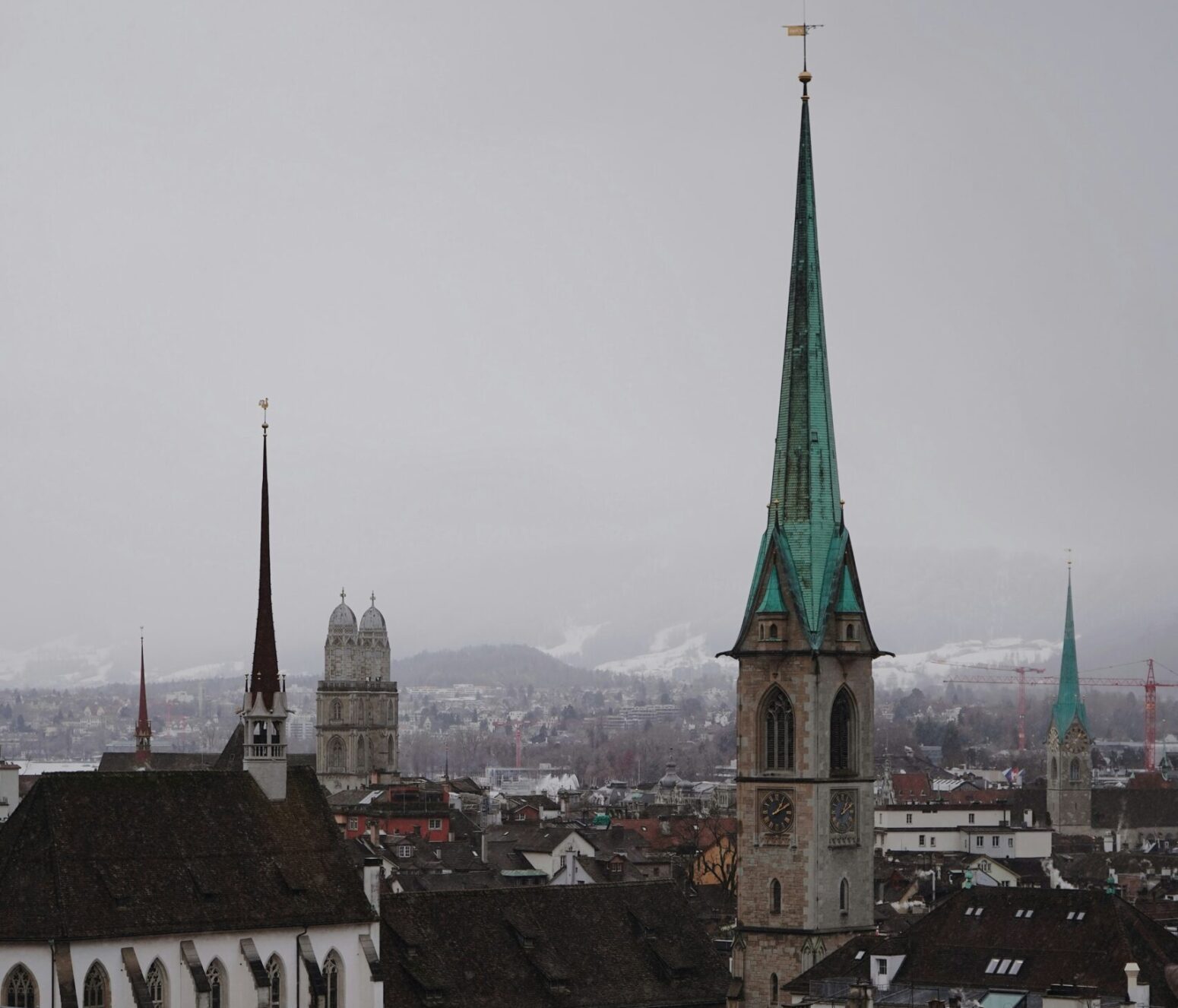 Winter view from a Zurich balcony. A few green and black-topped spires pierce a gray sky above the distant snow-capped Alps.