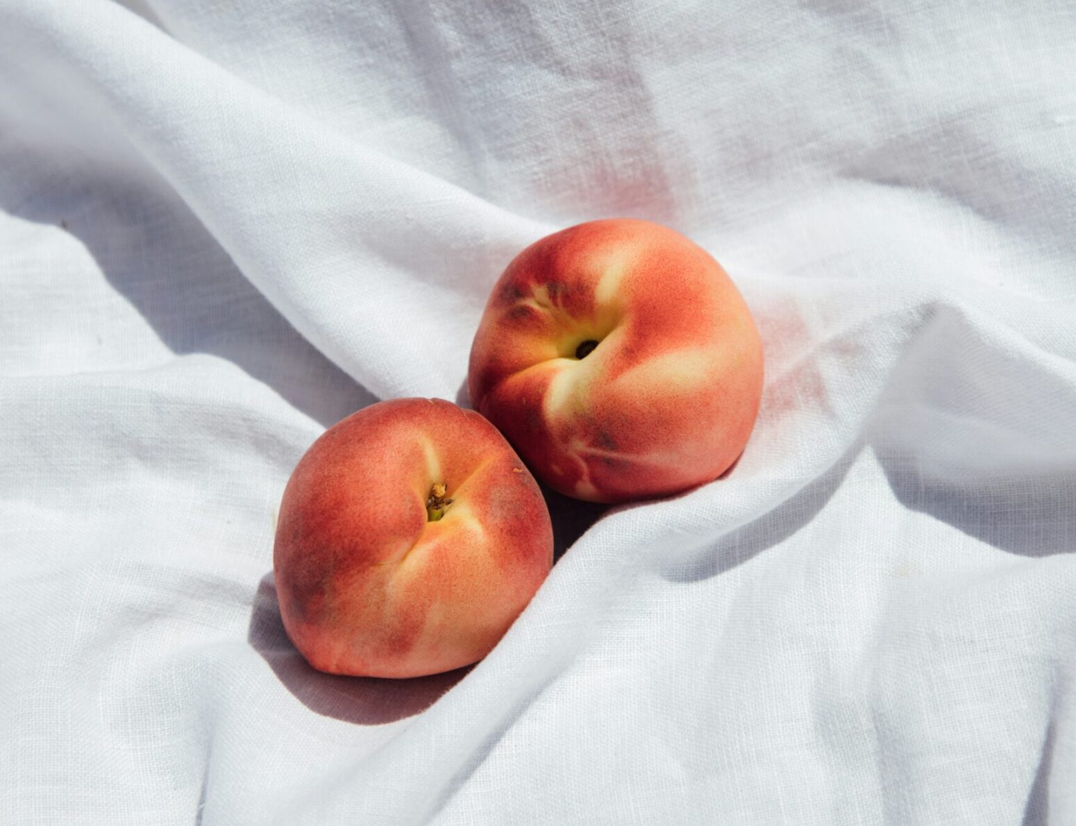 Two ripe peaches sit side-by-side on a white cloth, just touching.