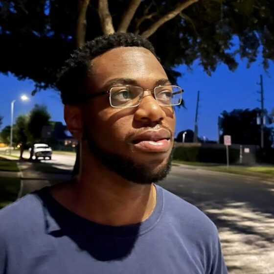 Kyle Okeke, a young Black man wearing rectangular glasses and sporting short hair and a beard, looks sideways. He stands on a wide street in the evening. The sky behind him is blue, as is his shirt. A car is parked on the street behind him.