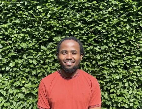 Migwi Mwangi, a Black man with a short beard, wears a red tee shirt in front of a wall of green leaves. He smiles broadly.