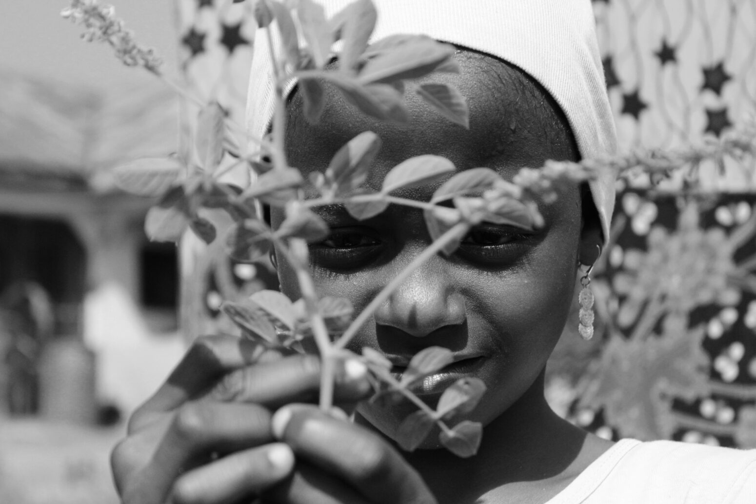 Black-and-white photo of a young Nigerian girl who holds a small branch in front of her face. She wears dangly earrings and a white head scarf.
