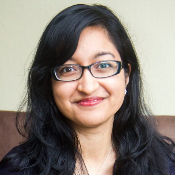 Woman smiling. Her hair is parted to the side, and she is also wearing glasses, red lipstick, and a black top.