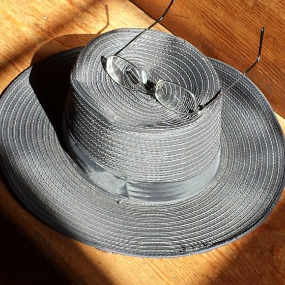 A gray straw-style hat on a wooden surface, with eyeglasses sitting on top, and sunlight.