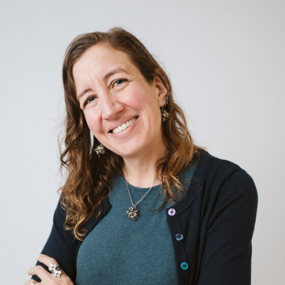 An Armenian American woman with light brown curly hair past her shoulders. She's leaning her head toward the left and smiling with crossed arms. She's wearing a black sweater, blue dress, and numerous colorful bracelets.
