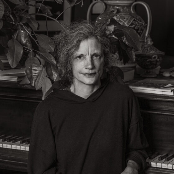 A black-and-white photo of a white woman in a dark sweater in front of a piano with ornate decor and a plant behind her.