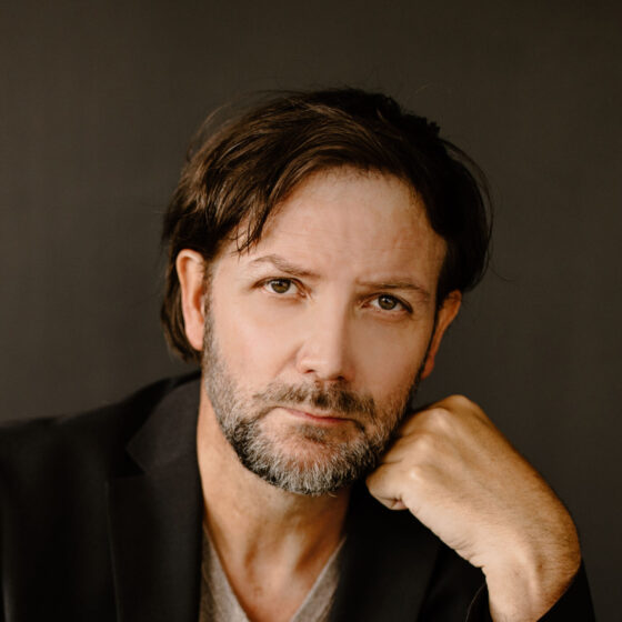 A white man with longish brown hair and a graying beard. He's leaning his face on his hand and wearing a black blazer over a gray T-shirt