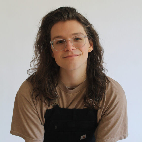 A white trans non-binary person with wavy hair and glasses. They are wearing a brown T-shirt and overalls.