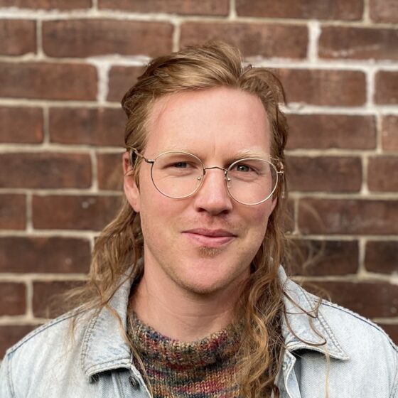 A white man with long auburn hair and glasses. He's wearing a jean jacket and standing in front of a brick wall