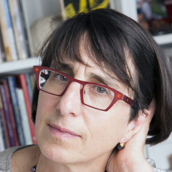 A woman with short brown hair and her hand behind her head. She's wearing red-rimmed glasses and is in front of bookshelves