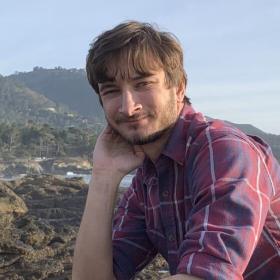 A white man with medium-length brown hair and a light beard, in a flannel shirt. He's sitting with his hand on his cheek, with hills and a bay in the distance