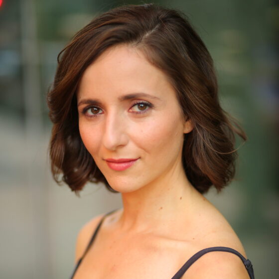 Danielle Frimer, a white Jewish woman with bobbed brown hair wearing a black tank top, stands in 3/4 profile. She smiles slightly.