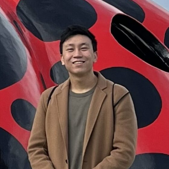 An Asian man in a brown blazer over a darker brown T-shirt, standing in front of an enormous red-and-black ladybug