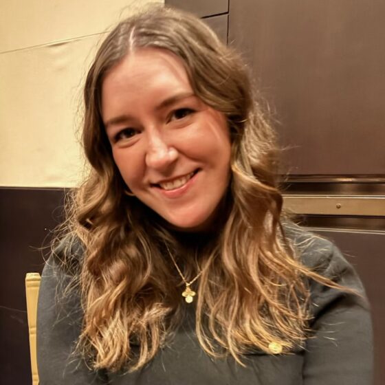 A white woman with long light-brown hair, smiling. She's wearing a gray shirt and gold necklace and standing in front of a divided wall
