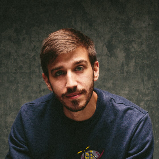A white man with brown hair and beard, wearing a dark blue shirt in front of a gray background. The image is mostly dark other than his face.