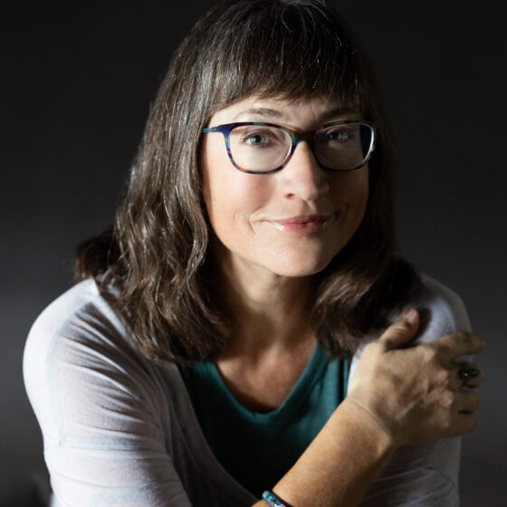 A white woman with bangs and glasses, smiling at the camera. She is wearing a grey cardigan.