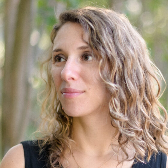 A woman with curly brown hair, wearing a black sleeveless shirt, with greenery behind her