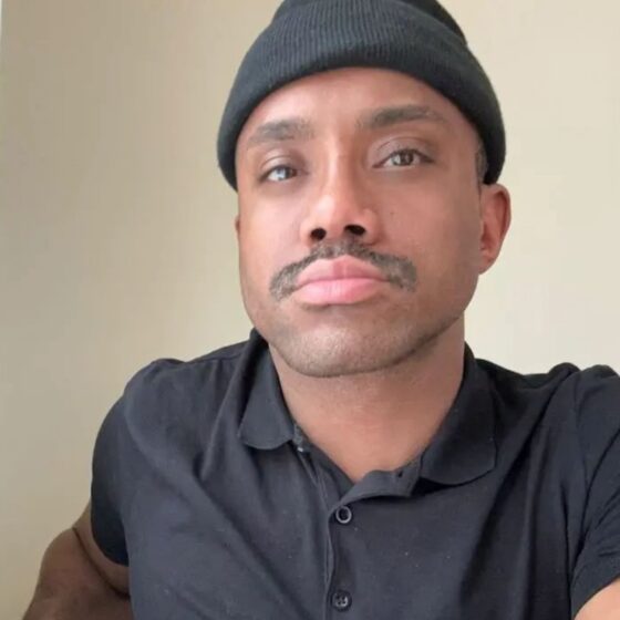 Kami Enzie, a multiracial man wearing a black beanie and polo, has a light mustache. He pouts slightly at the camera.