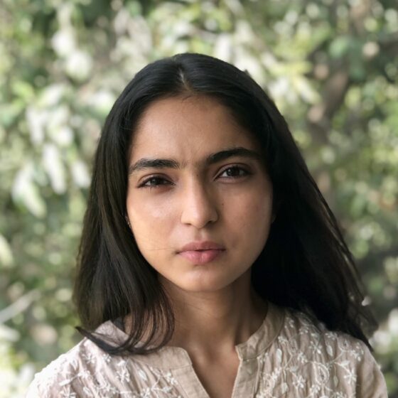 An Indian woman with long dark hair. She is wearing a tan textured cotton blouse and standing in front of greenery.