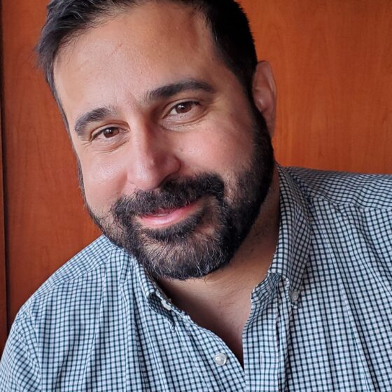A Cuban American man with a beard smiling for the camera. He is wearing a blue check shirt.