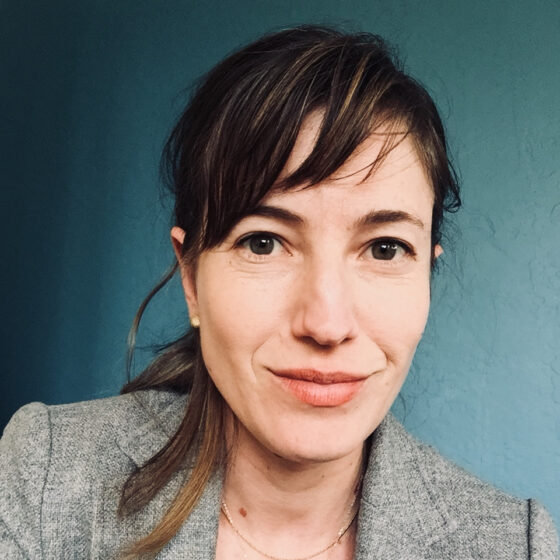 A headshot of a white woman with dark hair wearing a gray blazer and a necklace