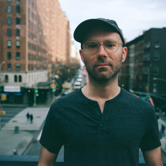 A white man with glasses and in a short-sleeved Henley shirt. He is backdropped by a city.