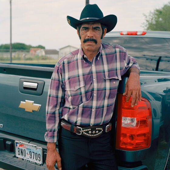 A latino man in cowboy hat, plaid shirt, belt, and black jeans, leans against the gate of his Chevy pickup. He has a dark, thick mustache and scowls.