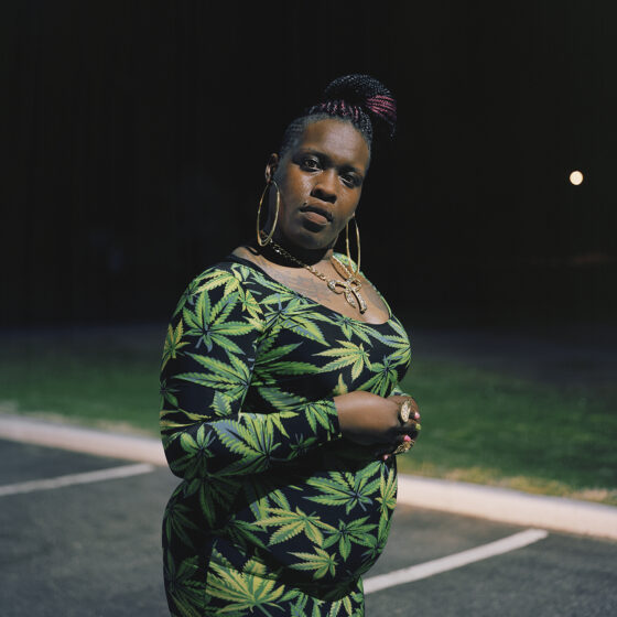 A Black woman wearing a tight-fitting dress with weed leaves printed on it, her braids pulled up into a neat twisted bun on top of her head, stands in an empty parking lot at night.
