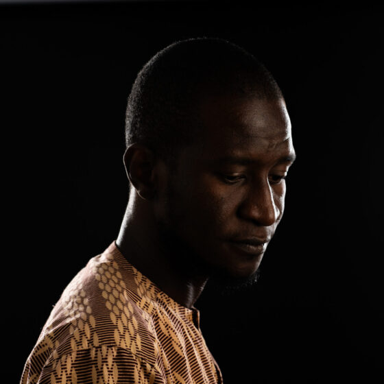 Saddiq Dzukogi, a Black African man, casts his gaze downward and is turned to us in 3/4 profile. He stands against a black backdroup and wears a yellow-and-green woven shirt. He is clean-shaven.