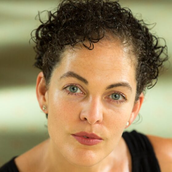 A woman with short dark curly hair and blue eyes. She's wearing stud earrings and a black sleeveless shirt