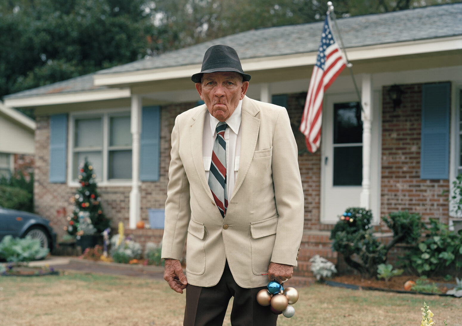 George, an old white man in fedora, tan jacket, brown pants, and red, white, and blue striped tie, holds some Christmas globes while scowling in his front yard. He's decorating. His pristine midcentury ranch home has blue shutters and an American flag hanging from a pole.
