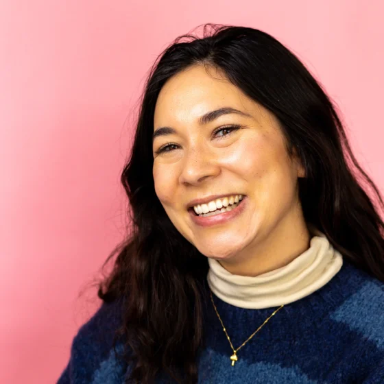 Natalie Villacorta, a mixed Asian American/white woman with long dark hair tossed across her shoulder, wears a blue and navy sweater over a turtleneck and a small gold necklace. She smiles broadly against a pink backdrop.