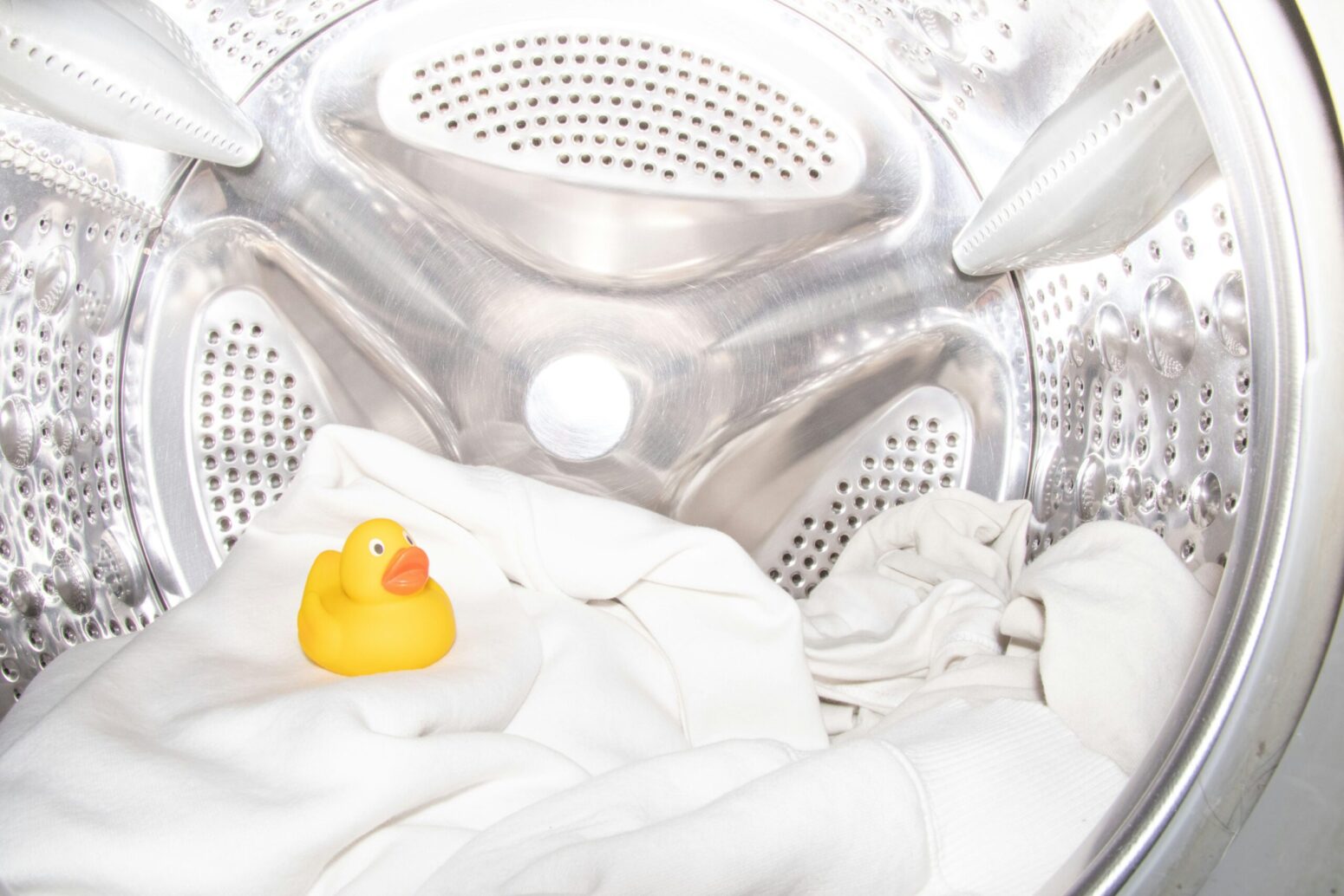 A pile of white fabrics topped with a yellow rubber duck sits in the reflective silver basin of a washing machine.