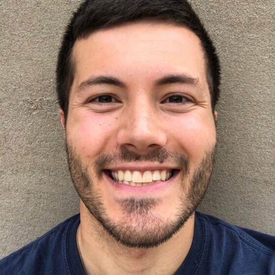 Matt Del Busto, a Latino man, bears a toothy smile. He has close-cropped dark hair and a five-o-clock-shadow. He wears a blue shirt and stands in front of an outdoor stucco wall.