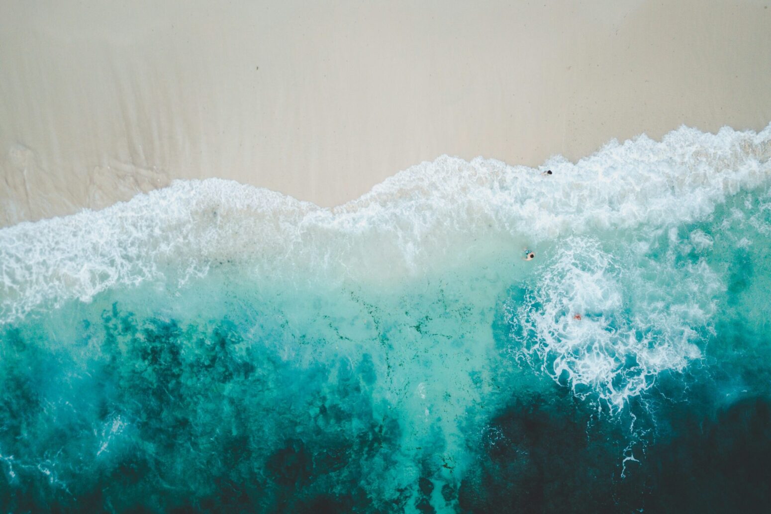 A beach from above, with dark waves becoming white and then tan sand above