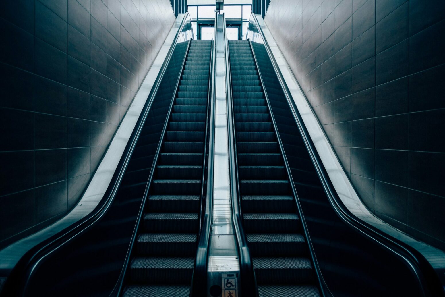 Two escalators side-by-side in a dark underground. A set of bright glass doors stands at the top of the escalator.